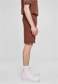 Bruine katoenen shorts met een elastische tailleband, een zijvak, gedragen met witte sneakers en sokken; zijaanzicht.