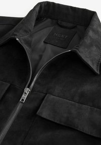 Schwarze Wildlederjacke mit einem Reißverschluss, ausgestattet mit einem strukturierten Kragen und zwei vorderen Taschen. Etikett im Inneren des Kragens sichtbar.
