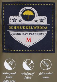 Námorná tkaninová nálepka s logom "Schmuddelwedda," so žltým a červeným textom, ikonou dažďa, ikonou odolnosti voči vetru a detailmi švov na svetlo hnedom pozadí.