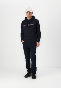 Navy hoodie with a front pocket, drawstring, and white and red horizontal stripe. Paired with dark jeans and black sneakers, complemented by a cap.
