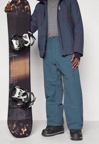 Osoba u plavim snježnim hlačama i tamnoj jakni drži crnu i smeđu dasku za snowboard s bijelim vezovima, stojeći na jednobojnoj pozadini.