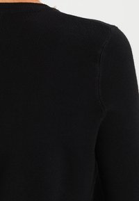 Svart ribbad tröja med rund halsringning, med en rad dekorativa knappar på baksidan. Mjuk textur och figursydd design.