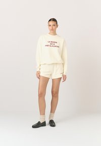 Kremowa bluza z czerwonym napisem, zestawiona z pasującymi kremowymi szortami, akcentowana czarnymi mokasynami i beżowymi skarpetkami, na jasnym tle.