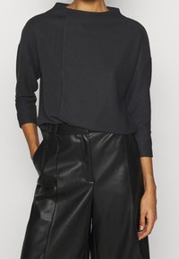 Maglione grigio scuro a coste con scollo a barca, caratterizzato da un pannello asimmetrico unico. Abbinato a pantaloni larghi in pelle nera.