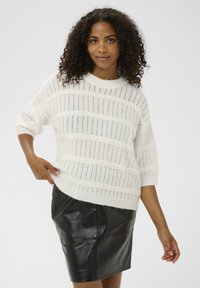 Femme aux cheveux bouclés portant un pull en tricot texturé blanc et une jupe en cuir noir, posant d'une main sur la hanche.