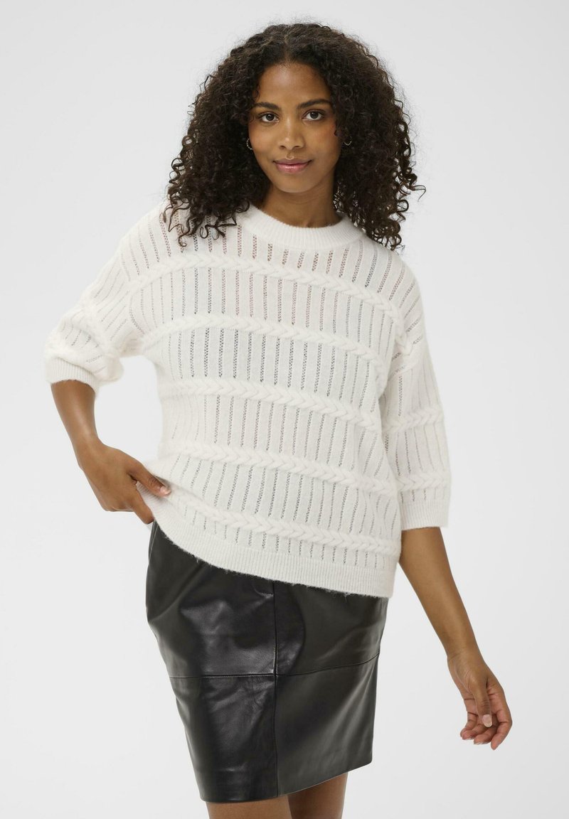 Femme aux cheveux bouclés portant un pull en tricot texturé blanc et une jupe en cuir noir, posant d'une main sur la hanche.