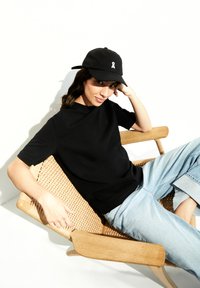 Zwarte T-shirt met korte mouwen, lichtblauwe jeans en een zwarte pet met een wit lintlogo. Zittend op een geweven houten stoel tegen een witte achtergrond.