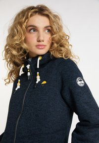 Marineblå hoodie med dragkedja fram, texturerat tyg och en gul paraply-logotyp. Har vita och gula snörningar med träpärlor.
