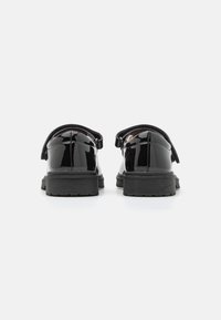 Zapatos de charol negro con punta redonda, correa ajustable y suela de goma gruesa con banda de rodadura texturizada. Vista trasera.