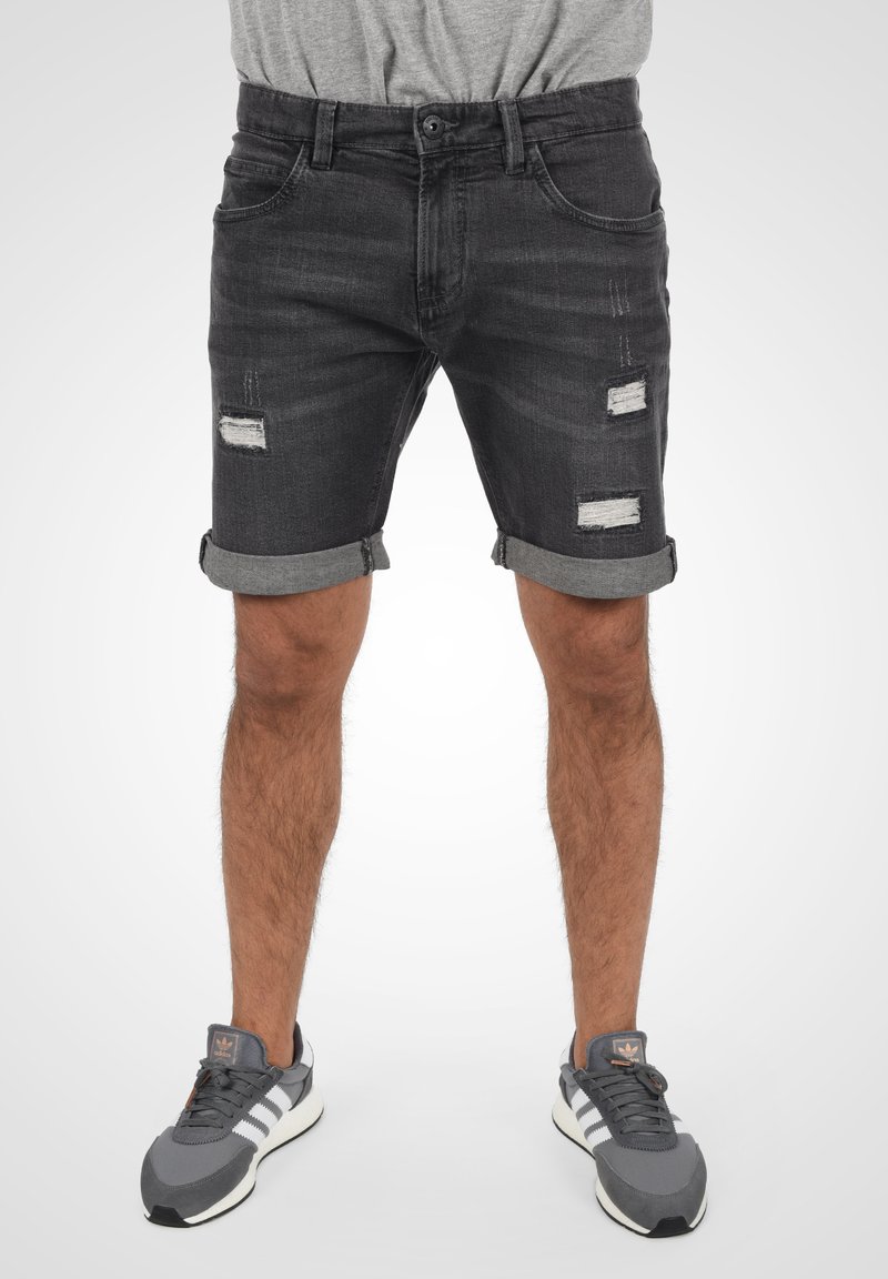 Schwarze Denim-Shorts mit umgeschlagenen Säumen; verfügt über verwaschene Stellen an der Vorderseite. Getragen mit grauen Sneakern. Glatte Textur.