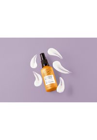 Reinigungsmilch für das Gesicht in einer orangen Flasche mit schwarzem Pumpspender, umgeben von weißen Cremeschwüngen, auf einem hellvioletten Hintergrund platziert.