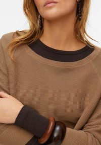 Bruine gebreide trui met een geribbelde halslijn en donkerbruine manchetten, gecombineerd met een houten armband. Focus op stof- en ontwerpd_details.