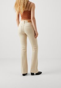 Mujer con cabello rizado que lleva pantalones acampanados beige, zapatos de tacón negros y un top corto con estampado naranja, de pie frente a un fondo blanco liso.