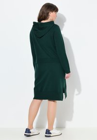 Abito verde lavorato a maglia con cappuccio, maniche lunghe, polsini e orlo a costine, con spacchi laterali, abbinato a sneakers bianche con accenti blu.