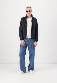 Jachetă neagră ușoară cu guler înalt, pulover crem dedesubt și pantaloni cargo din denim albastru, largi. Adidașii completează ținuta.