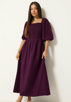 Donna con capelli lunghi e lisci che indossa un vestito maxi viola con maniche a sbuffo e ricami floreali sul corpetto, in piedi davanti a un muro semplice.