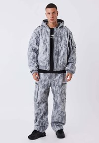 Grijze tie-dye jas met capuchon en elastische boorden, gecombineerd met bijpassende cargobroek met ritsen en meerdere zakken. Zwarte sneakers.