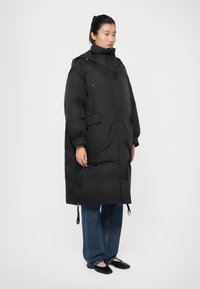 Doudoune noire avec un col montant, fermeture éclair à l'avant, deux poches latérales et des détails à cordon. Portée sur un jean bleu à jambes larges et des ballerines noires.