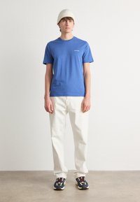 Blue cotton t-shirt with a round neck and subtle logo, paired with white denim pants. The model wears a ribbed beige beanie and multicolored sneakers.