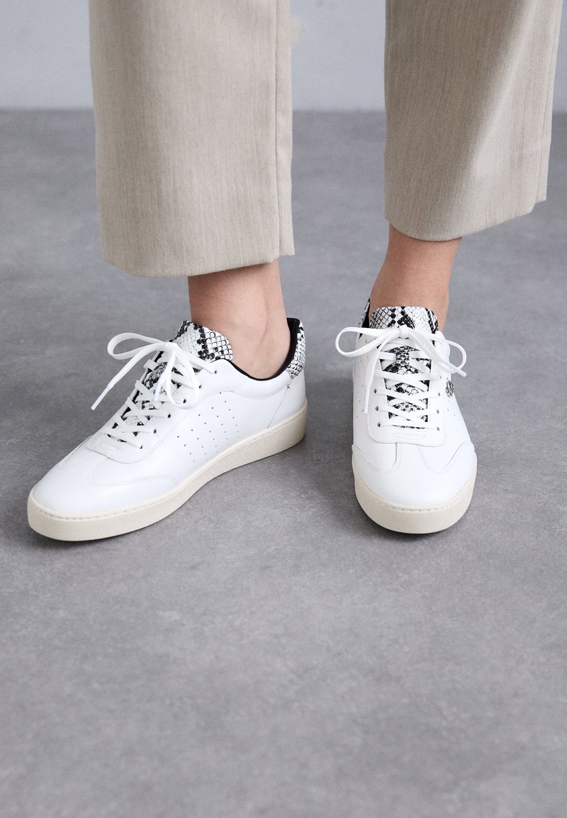 Sneakers bianche in pelle con perforazioni, colletto a motivi bianco e nero e suola in gomma testurizzata. Abbinati a pantaloni grigi chiari.