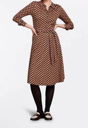 Mujer con vestido abotonado hasta la rodilla con un patrón geométrico rojo y beige, medias negras y mocasines blancos y negros, con las manos en las caderas.