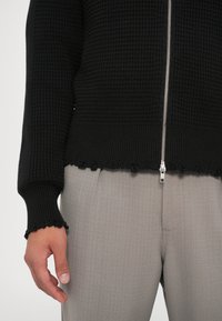 Černý pletený cardigan s texturovaným vzorem, stříbrným zipem a vlnkovaným lemem na výstřihu a manžetách, kombinovaný se světle šedými kalhotami.