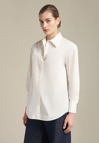 Witte blouse met kraag, lange mouwen en een afgeronde zoom. Gemaakt van een zachte stof met een subtiele glans en knopen aan de voorkant.