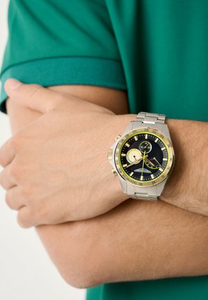 Orologio in acciaio inossidabile con finitura argento e dorata, quadrante nero, molteplici funzioni cronografiche e dettagli verdi sulla lunetta e sulle lancette.