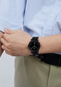 Reloj analógico negro con una esfera redonda, marcadores de horas blancos, detalles rojos y una correa de cuero negra, llevado en una muñeca. Camisa a rayas azul visible.