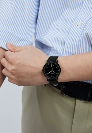 Montre analogique noire avec un cadran rond, des marqueurs d'heure blancs, des accents rouges et un bracelet en cuir noir, portée au poignet. Chemise rayée bleue visible.