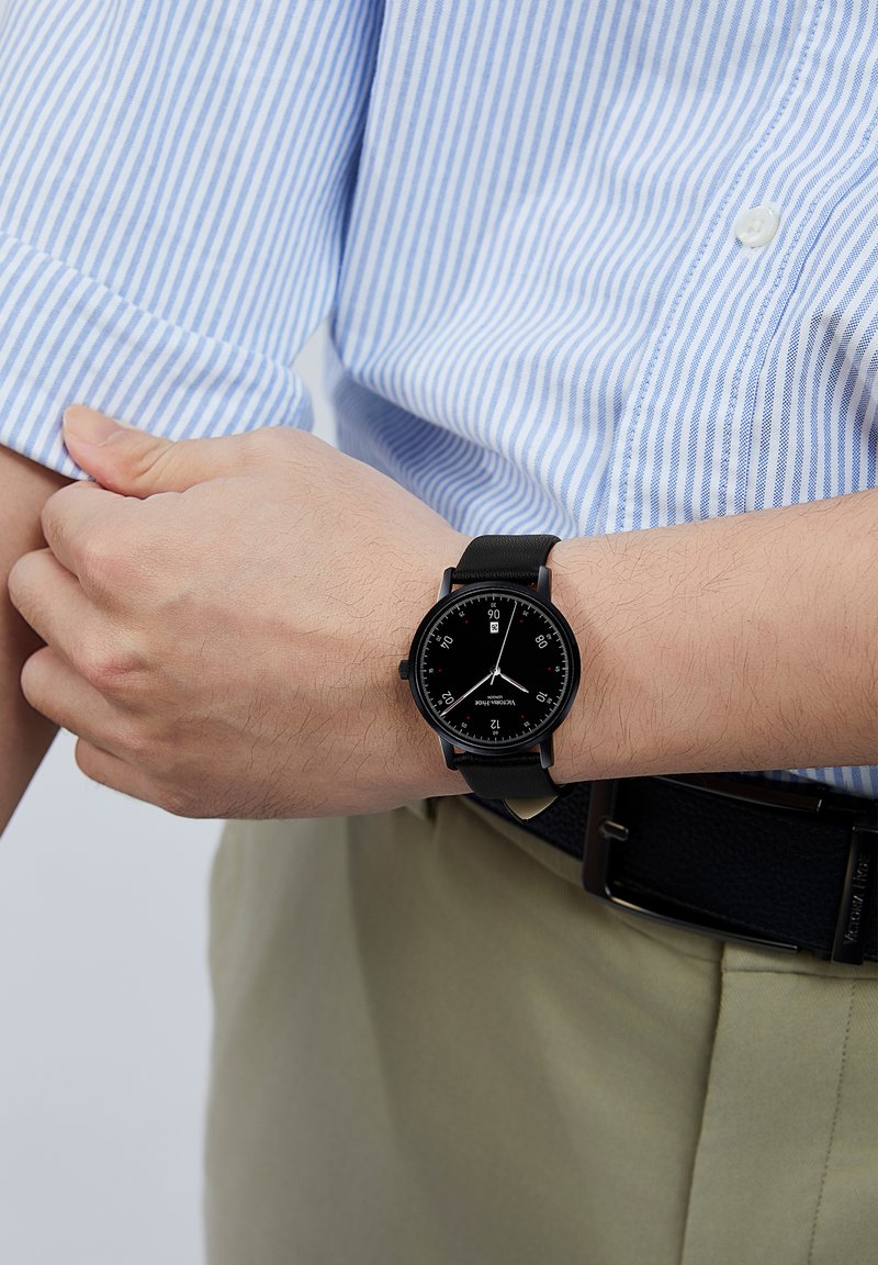 Reloj analógico negro con una esfera redonda, marcadores de horas blancos, detalles rojos y una correa de cuero negra, llevado en una muñeca. Camisa a rayas azul visible.