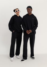 Deux individus portent des sweatshirts à capuche oversize noirs assortis et des pantalons. L'un porte des lunettes de soleil blanches ; l'autre porte une casquette noire. Les chaussures sont de couleurs variées.