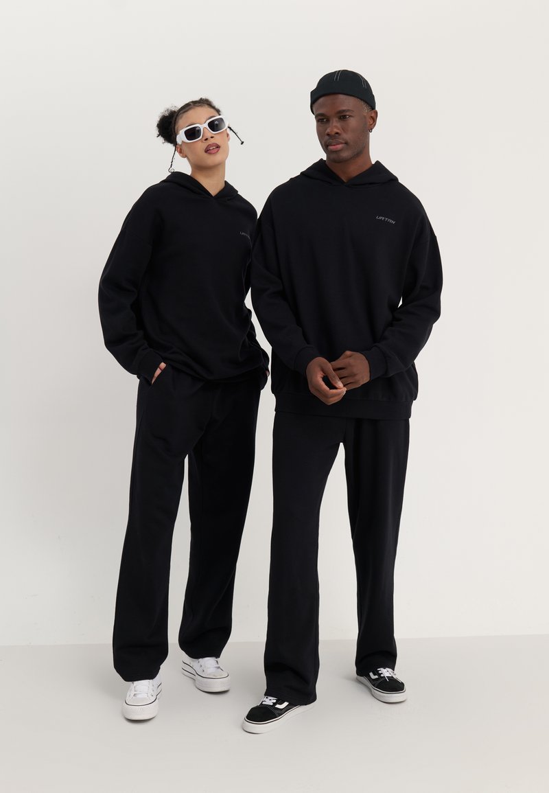 Deux individus portent des sweatshirts à capuche oversize noirs assortis et des pantalons. L'un porte des lunettes de soleil blanches ; l'autre porte une casquette noire. Les chaussures sont de couleurs variées.