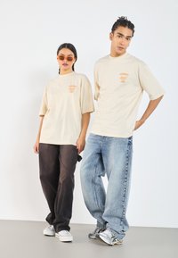 Beige graphic t-shirts with "YUMMY" print in orange, paired with dark brown and light blue baggy jeans. White sneakers complete the outfits.