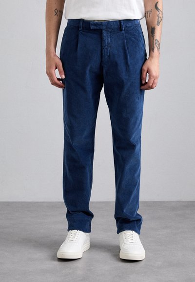 Pantalons en velours côtelé bleu avec une coupe droite, présentant des plis sur le devant et un tissu texturé. Portés avec des sneakers blanches.