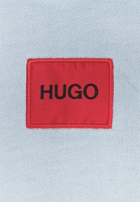 Tejido de punto azul claro con un prominente parche rectangular rojo que presenta el texto bordado en negro "HUGO" centrado en él.
