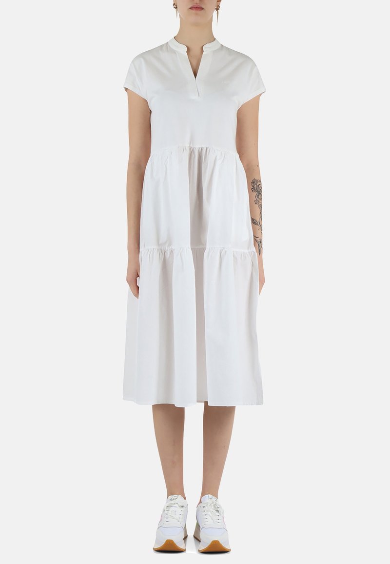 Abito bianco in cotone con colletto corto, maniche corte e gonne a strati. Indossato con sneakers bianche, evidenziando uno stile informale.