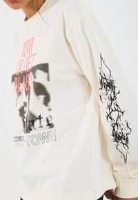 Long-sleeve cream cotton t-shirt featuring bold, graphic text in black and pink, with abstract black artwork on the right sleeve.