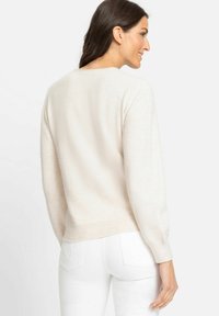 Helle beige Strickjacke mit weicher Textur, rundem Ausschnitt und geripptem Saum. Sie hat lange Ärmel und einen lässigen Schnitt. Weiße Hosen sind sichtbar.