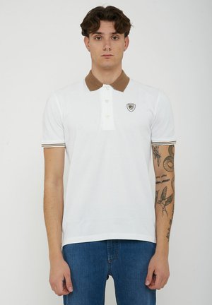 Young man with curly hair wearing a white polo shirt with brown collar and sleeve trim, blue jeans, tattoos visible on right arm.