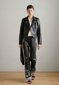 Veste de moto en cuir noir, haut blanc, pantalon évasé gris avec broderies florales, chaussures noires à lanières, tenant un sac noir.