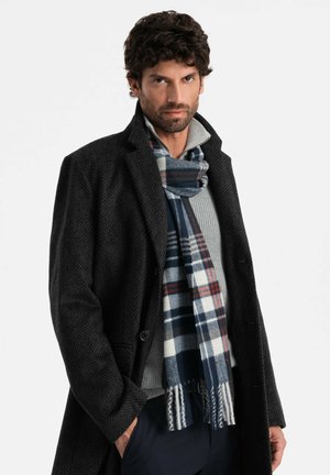 Abrigo negro texturizado sobre un suéter gris, combinado con una bufanda a cuadros azul y blanco con acentos rojos. Modelo posa con las manos en los bolsillos.