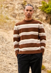 Maglione a righe marrone e crema con collo tondo e polsini a coste, abbinato a pantaloni scuri, su uno sfondo roccioso.