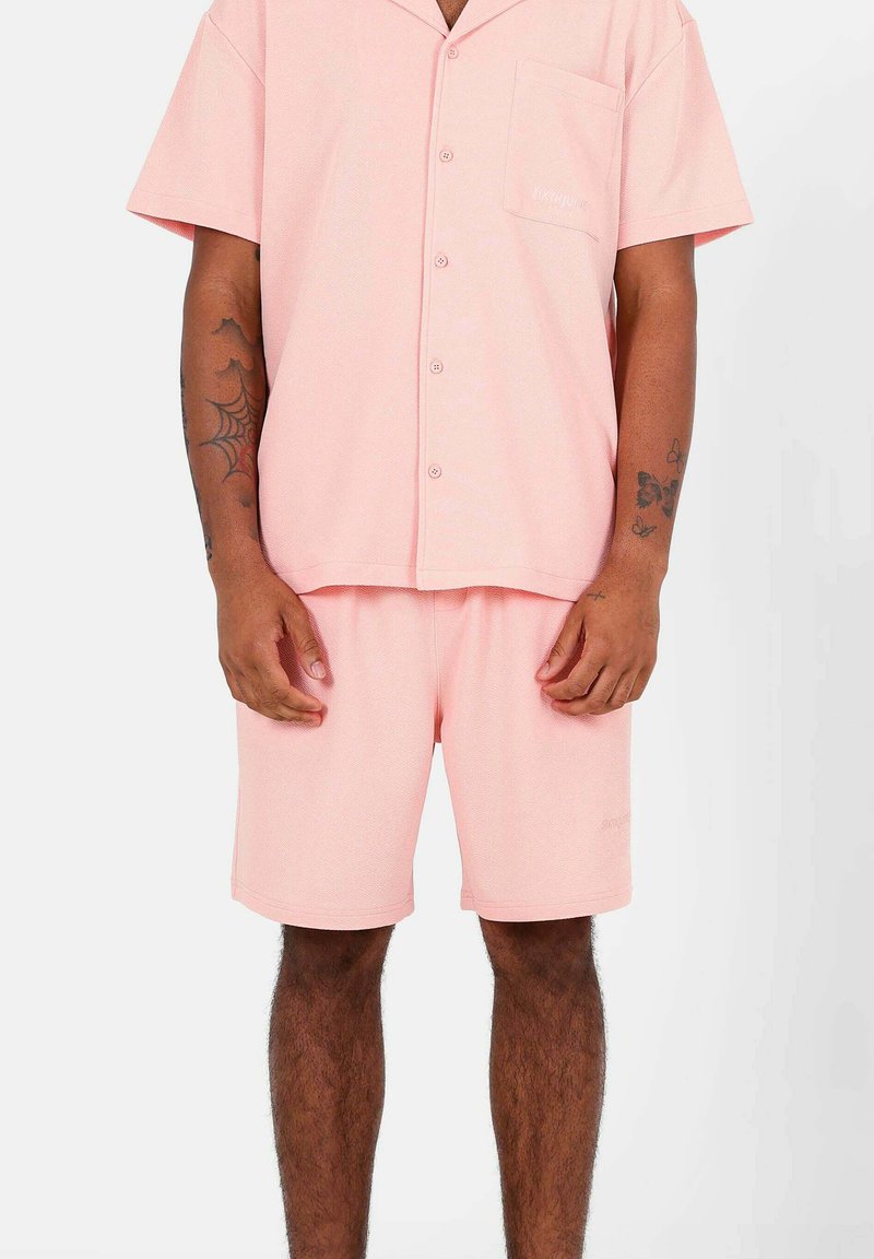 Camicia a maniche corte button-up rosa chiaro con tasca, abbinata a pantaloni corti coordinati. Il tessuto ha una finitura testurizzata e una vestibilità comoda.