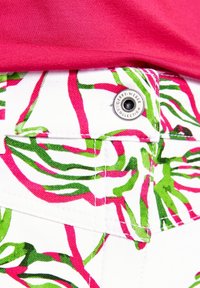 Pantalons blancs avec un motif floral rose et vert. Présente des accessoires en métal avec le nom de la marque sur la taille. Tissu texturé.