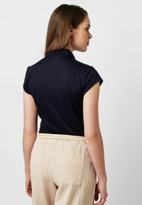 Marineblaues Poloshirt mit kurzen Ärmeln und Kragen, glatte Textur, kombiniert mit hellbeigen Hosen mit geflochtenem Taillenbund und zwei Gesäßtaschen.
