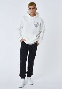 Sudadera blanca con capucha y diseño gráfico gris, con bolsillo frontal, combinada con pantalones cargo negros que tienen varios bolsillos, y zapatillas blancas.