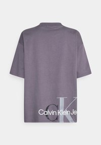 Camiseta gris de gran tamaño con cuello redondo, mangas cortas y el logotipo "Calvin Klein Jeans" impreso en blanco y negro en el lado inferior.