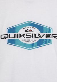 Bílý tričko s velkým hexagonálním logem Quiksilver v modré a černé barvě, s nápisem "The Original Boardriding Company" nahoře.