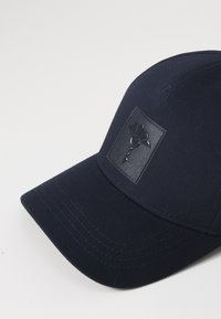 Sötétkék baseball sapka textilből, textúrázott fekete logóval az elején, ívelt napellenzővel és varrott részletekkel.